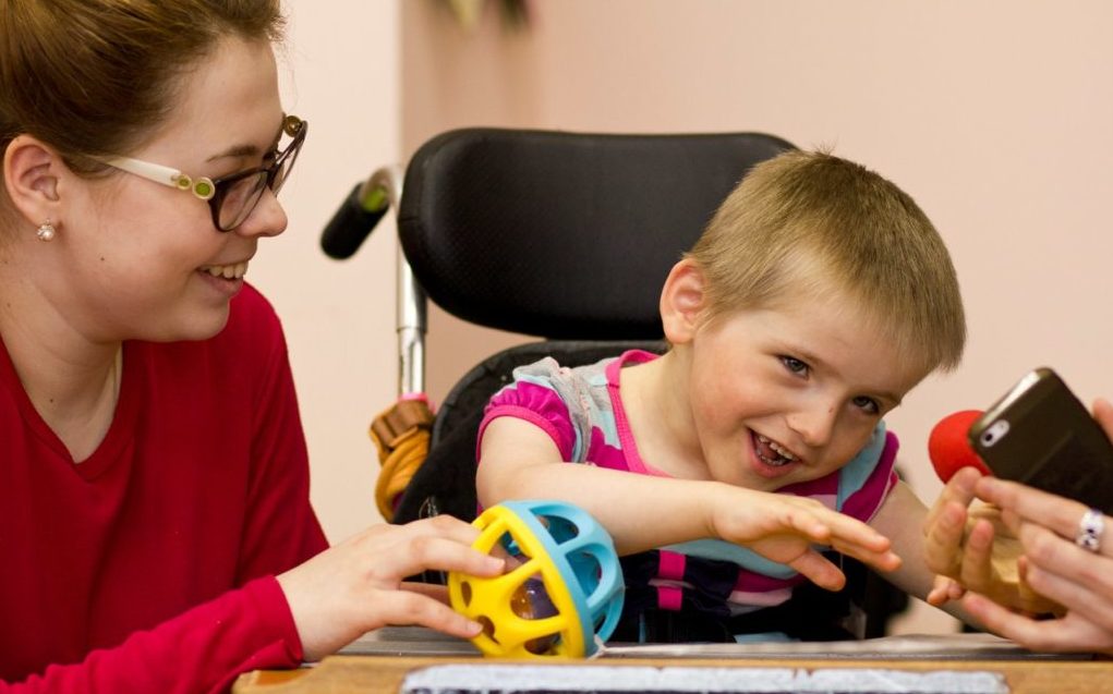 Ein Kind mit eingeschränkten motorischen Fähigkeiten sitzt in einem Rollstuhl an einem Tisch – es lacht. Neben ihm sitzt eine Betreuerin und hält ihm einen bunten Ball hin.