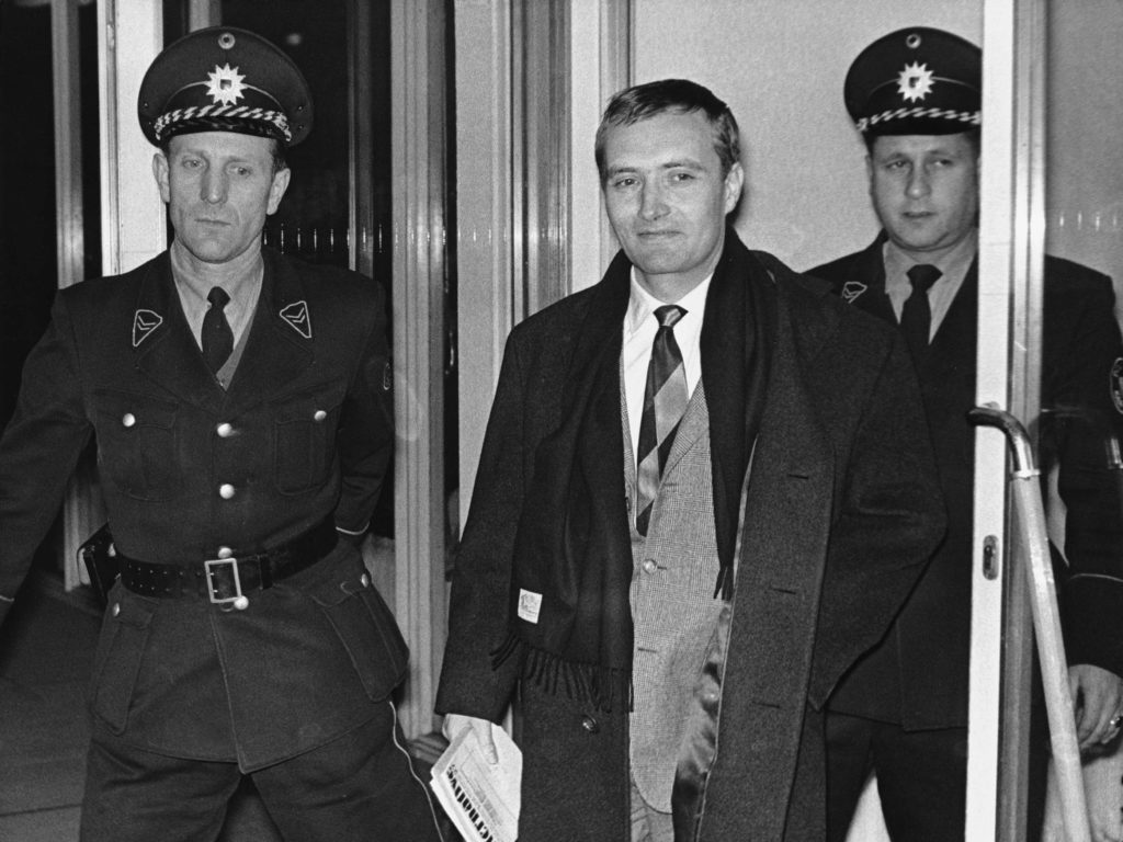 Über uns > Fotografie der Festnahme Rudolf Augsteins im Jahr 1962. Der Journalist tritt – begleitet von zwei uniformierten Beamten – lächelnd aus einer Tür.